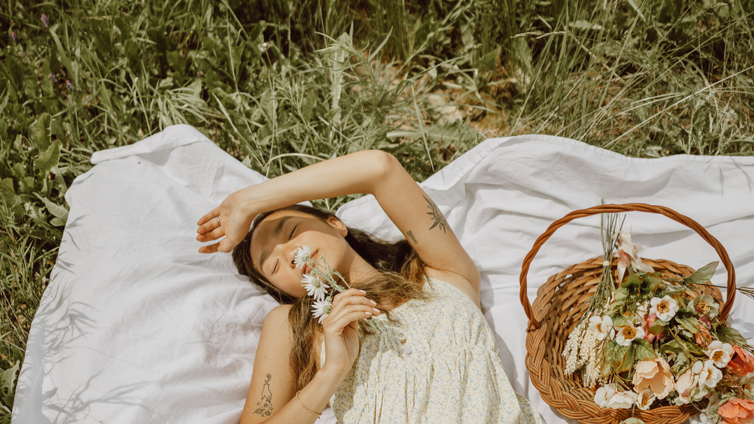 Person smelling a flower while lying on a white blanket next to a basket of flowers.
