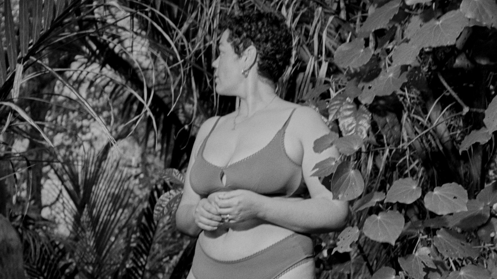 Black and white image of person wearing AWWA facing away from the camera, surrounded by palms and kawakawa